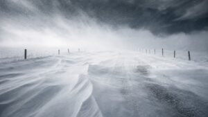 Severe blizzard blowing across an open rural road with deep drifting snow and near-whiteout visibility.